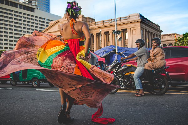 Visual of a protestor in rainbow fabric across the street from the Presidential Secretariat as a couple on a motorbike look on.